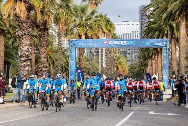 People dressed in blue ride bicycles down a road under a start banner that says MACA Cancer 200 Ride for the Perkins